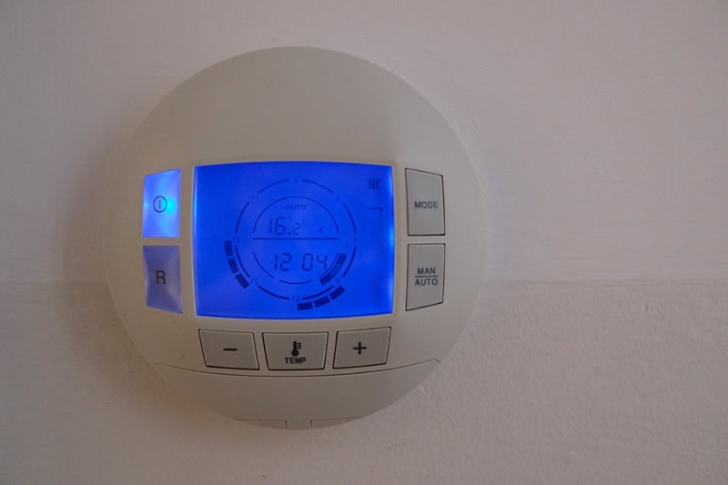 How to Set Programmable Thermostat. Photo of a digital thermostat mounted on a wall with a blue light.
